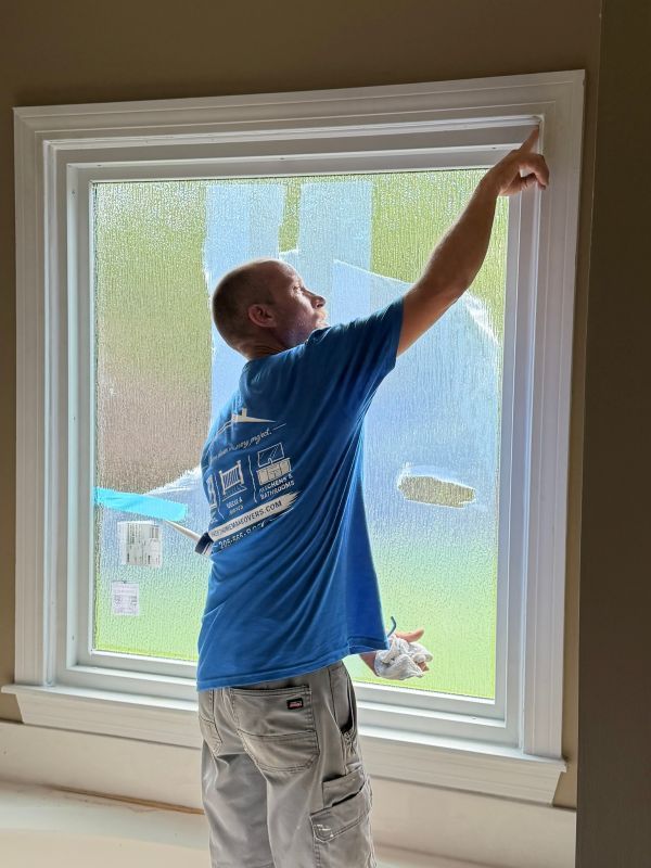 An installer in a blue Sweet Home Makeovers T-shirt works on a large new window with white interior trim.