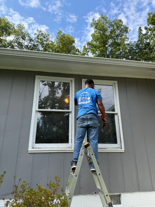 An installer in a blue T-shirt stands on a ladder, working on a window of a gray-sided home.